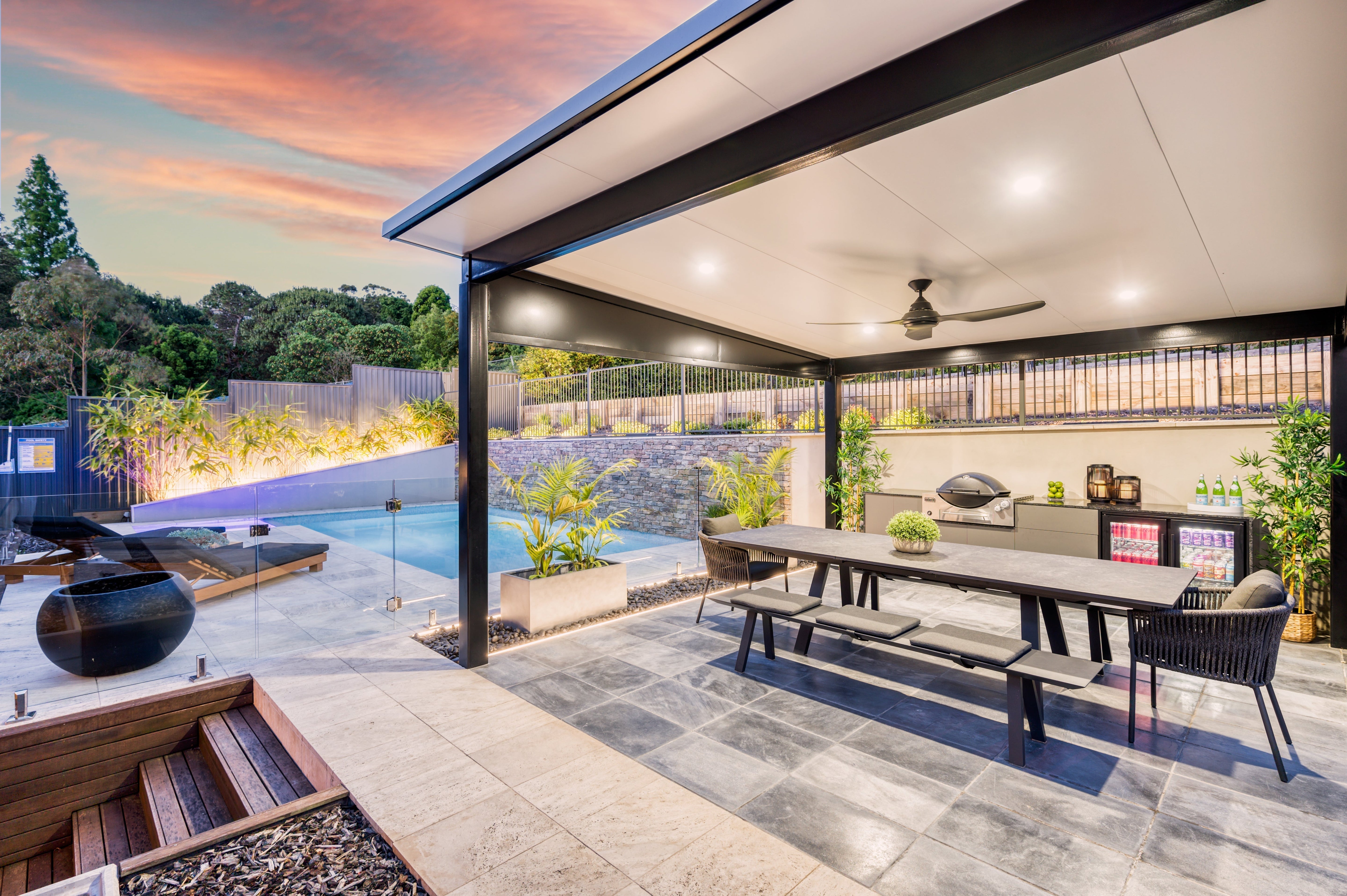 Modern outdoor entertaining area with Versiclad insulated roof, integrated lighting, dining table, outdoor kitchen, and poolside retreat surrounded by greenery.